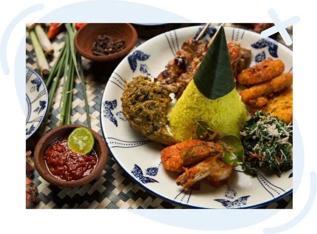 Plate of Indonesian cuisine featuring a cone of yellow turmeric rice surrounded by assorted side dishes, chili sambal, prawns, vegetables, and fried items, presented on a patterned plate with small bowls of condiments nearby.