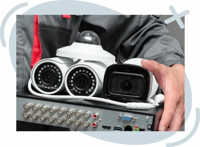 Close-up of various security cameras resting on a DVR unit held by a technician in a gray work jacket.