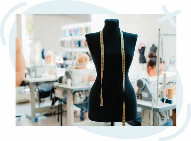 Black dress form with a yellow measuring tape draped around its neck in a bright tailoring studio with people working at sewing machines in the background.