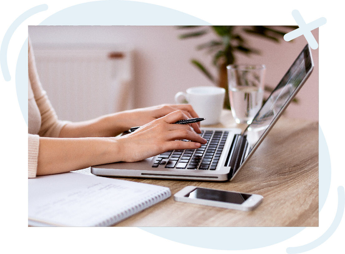 Close-up of hands typing on a laptop at a tidy desk with a notebook, smartphone, and a cup and glass in the background.