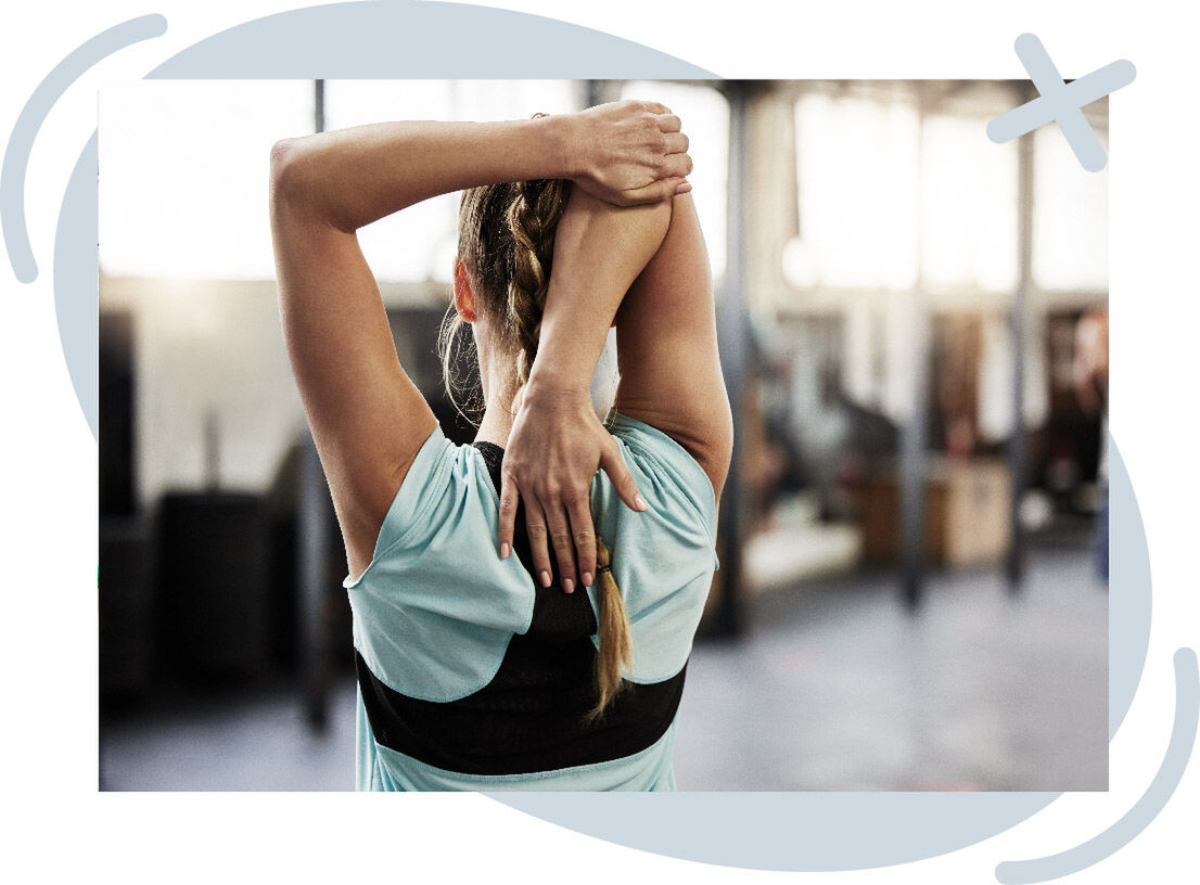 Person stretching their triceps by reaching one arm overhead and the other hand up the back, seen from behind in a gym setting.