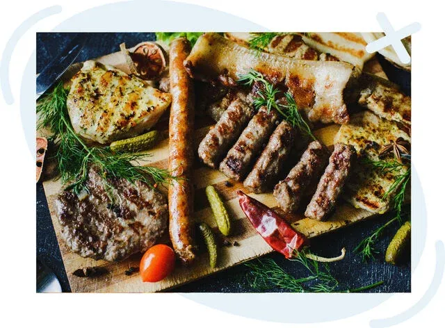 Assorted grilled meats and sides on a wooden board, including sausages, burger-style patties, grilled cheese slices, pickles, herbs, and a dried chili pepper.