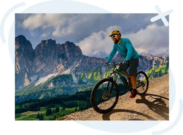 Mountain biker riding up a dirt trail with dramatic rocky peaks and green valleys in the background under a cloudy sky.
