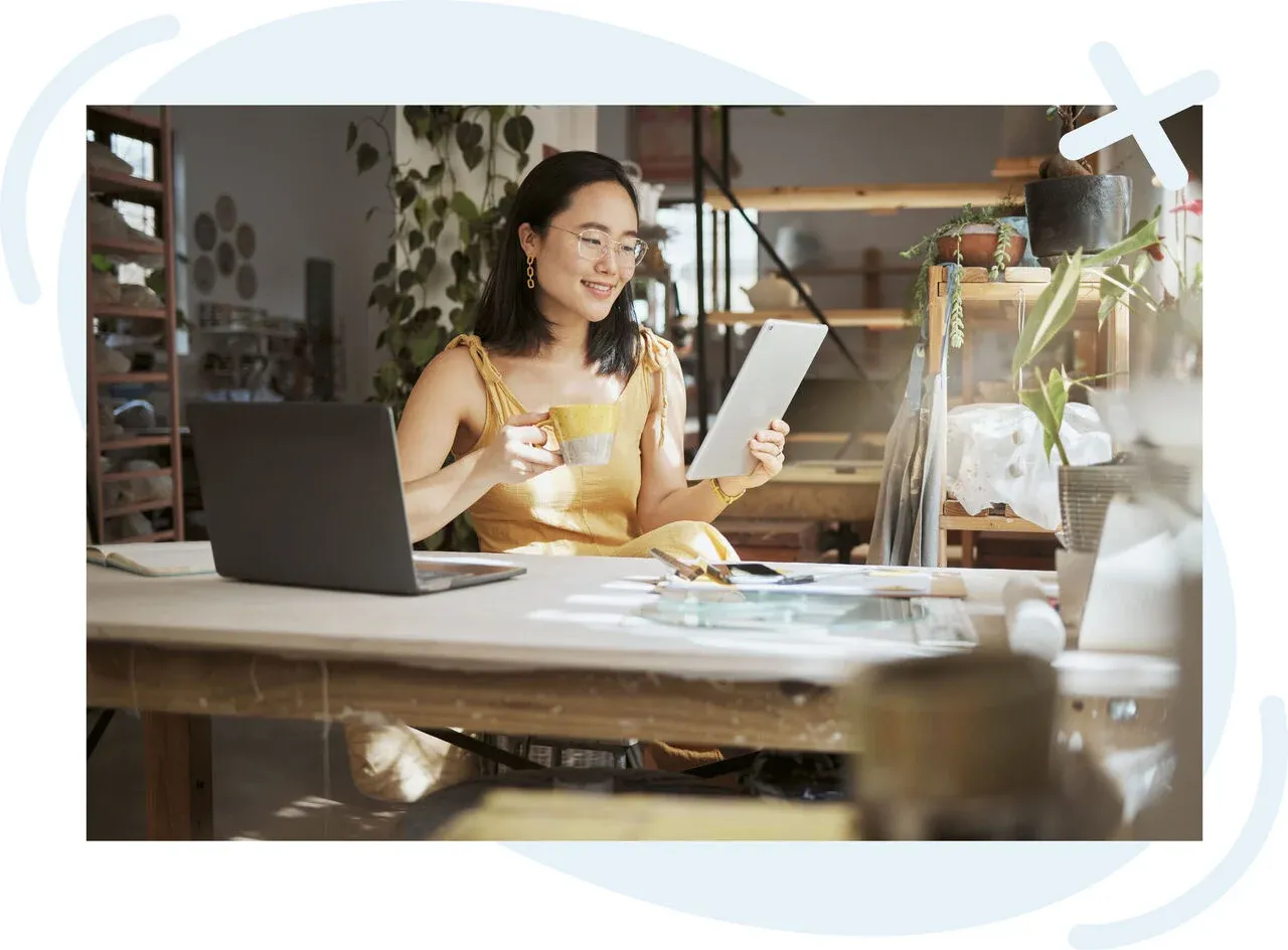 Person in a sunlit studio working at a large table, smiling while holding a tablet and a yellow mug, with a laptop open nearby and plants on shelves in the background.