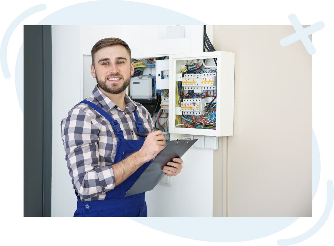 Electrician inspecting an open electrical panel while holding a clipboard and pen, smiling at the camera.