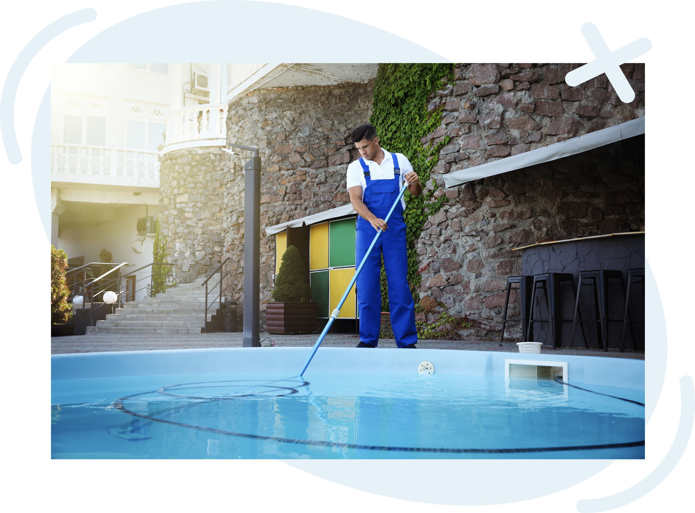 Worker in blue overalls cleaning a swimming pool with a long pole near a stone-walled terrace.
