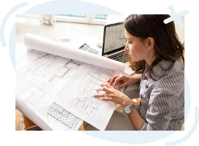 Person reviewing architectural blueprints at a desk with a laptop and calculator nearby.