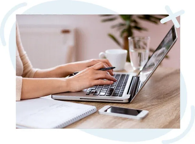 Close-up of a person typing on a laptop at a wooden desk with a pen in hand, notebook, smartphone, coffee cup, and glass of water nearby in a softly lit home office setting.