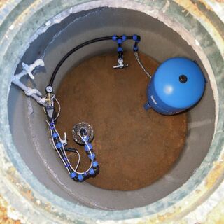Top-down view into a circular underground utility pit with plumbing and a blue pressure tank installed inside.