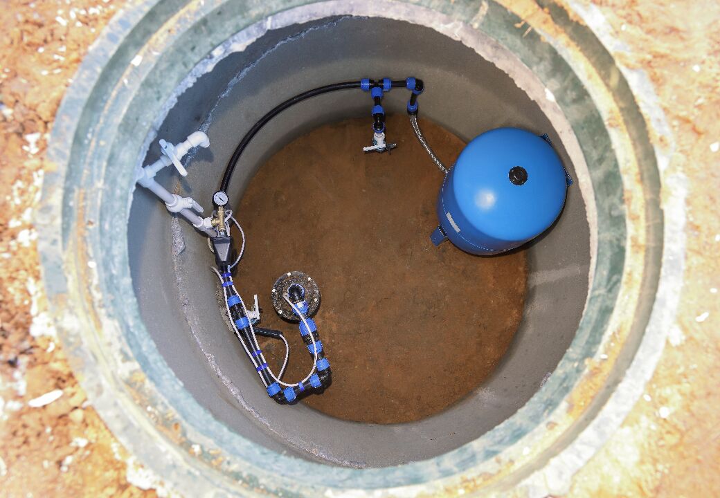 Top-down view into a circular underground utility pit with plumbing and a blue pressure tank installed inside.