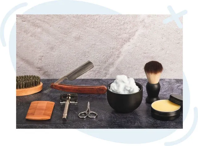 Assorted traditional barber tools laid out on a dark countertop, including a straight razor, shaving brush, bowl of foam, scissors, comb, safety razor, beard brush, and tin of balm.