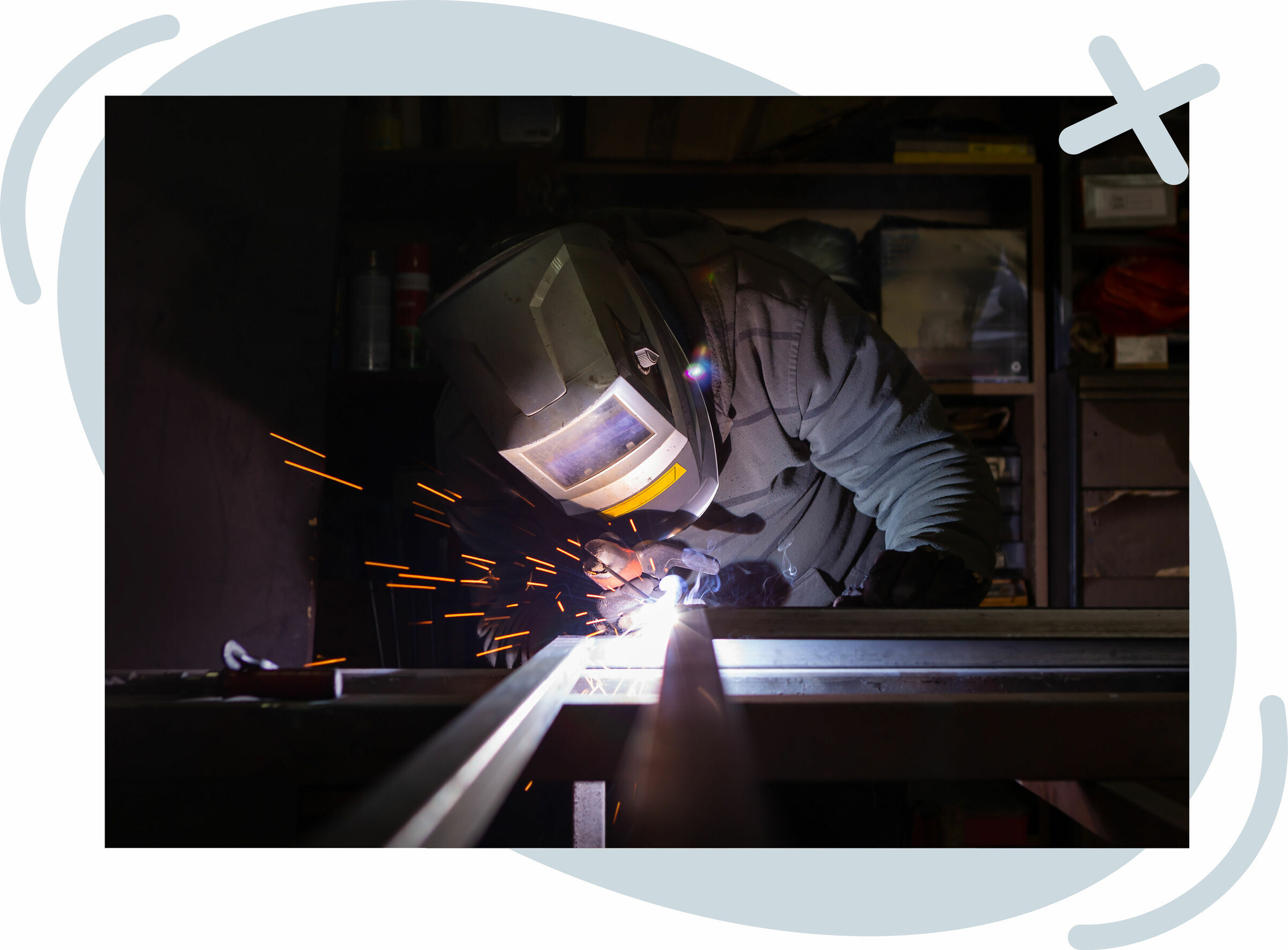 Person wearing a welding helmet and protective clothing welds metal in a dim workshop, with bright sparks and a bluish arc lighting their hands and the workpiece.