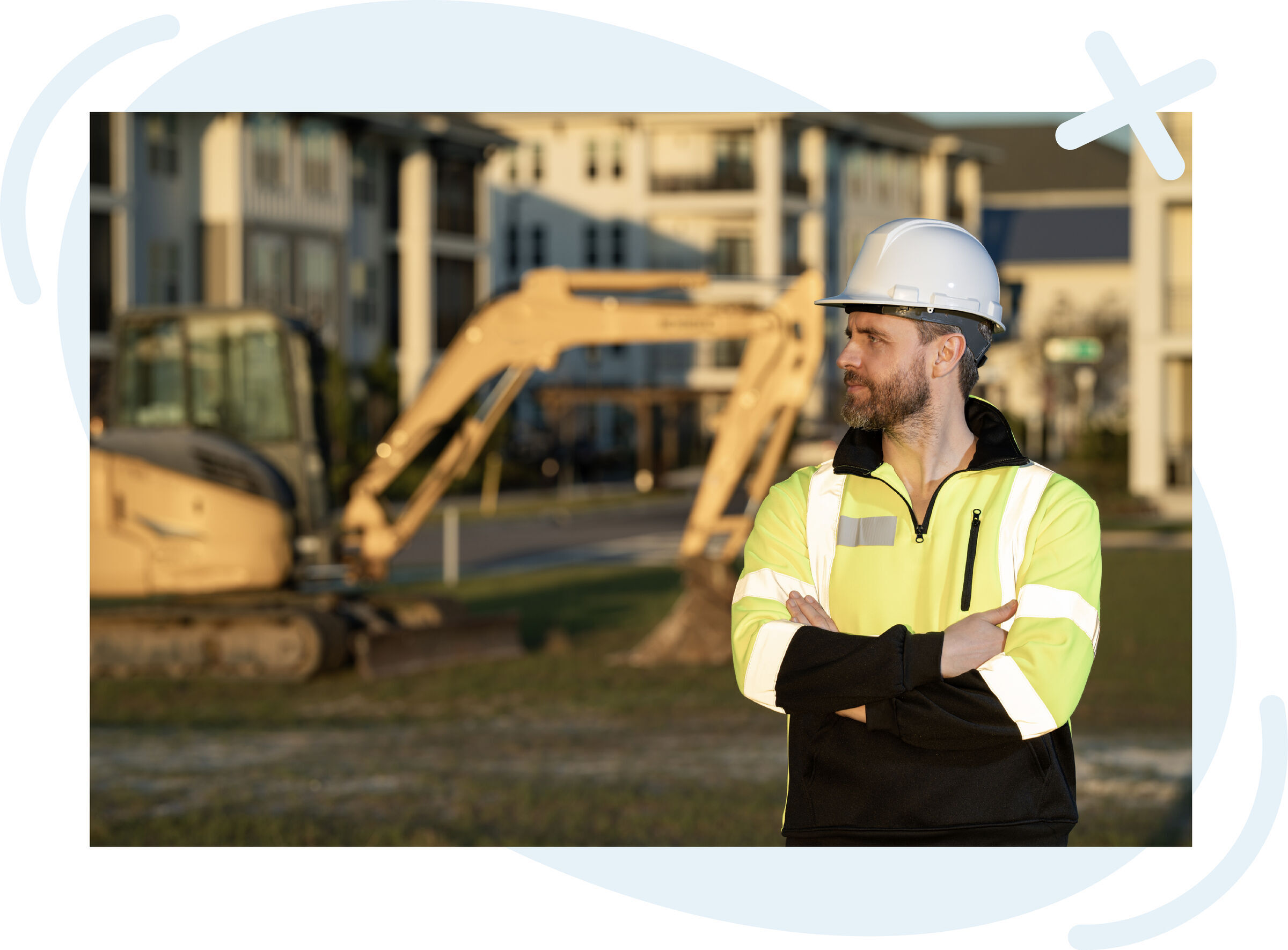 Construction worker in a neon safety jacket and white hard hat standing with arms crossed at a job site, with a blurred excavator and apartment buildings in the background at sunset.