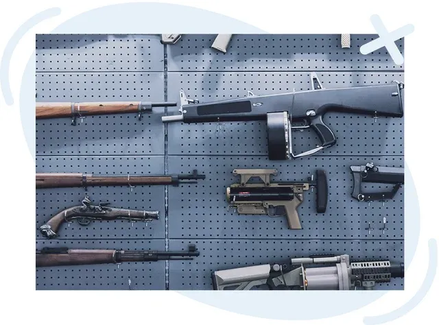 Display wall with various firearms mounted on a gray perforated panel, including rifles, a compact submachine gun, a large drum‑magazine weapon, and firearm components arranged in rows.