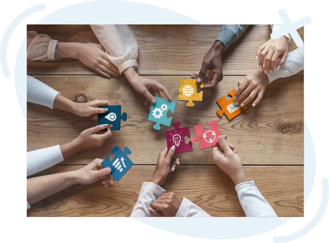 Top-down view of a diverse group of people around a wooden table, each hand holding a colorful puzzle piece with business-related icons, bringing the pieces together in the center to symbolize teamwork and collaboration.