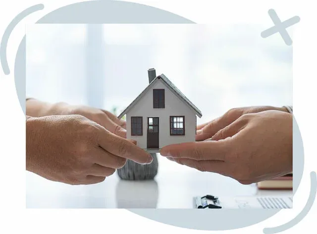 Two pairs of hands gently holding a small model house together, suggesting a real estate agreement or home purchase. Soft, bright lighting with a blurred office-like background and stationery items on a table.