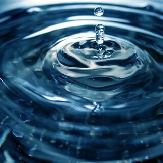 Close-up of a water droplet splashing into a calm surface, creating ripples and a small column of water with a second droplet above it.