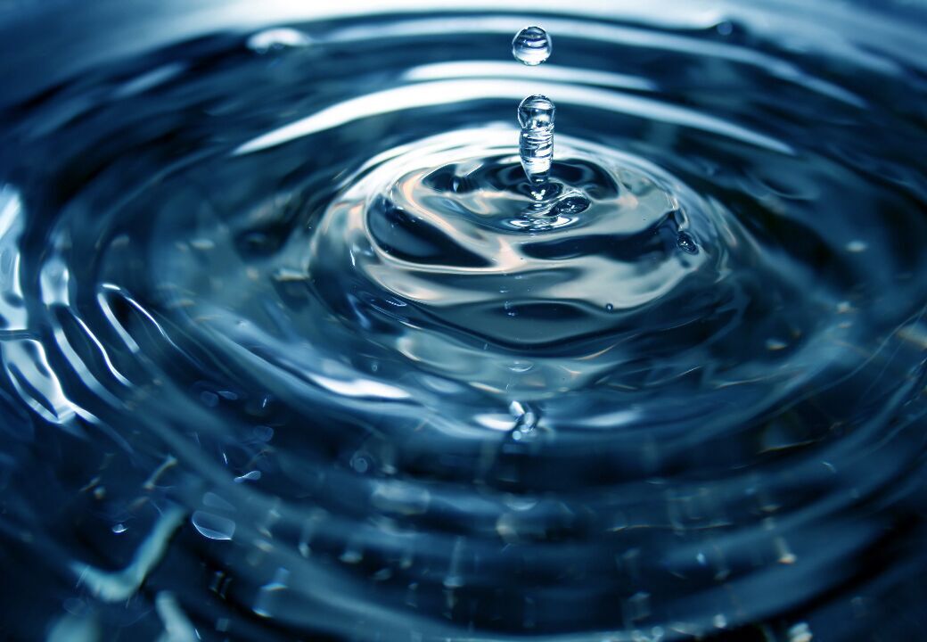 Close-up of a water droplet splashing into a calm surface, creating ripples and a small column of water with a second droplet above it.
