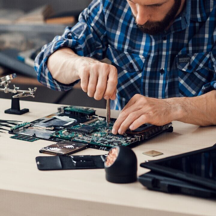 Person in a blue plaid shirt repairing a laptop motherboard on a workbench with precision tools and electronics parts spread out.
