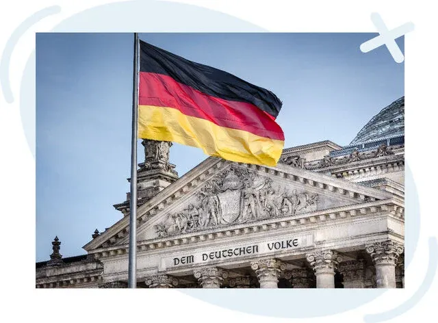 German flag waving in front of a grand stone building with classical columns and ornate pediment, cloudy sky in background.