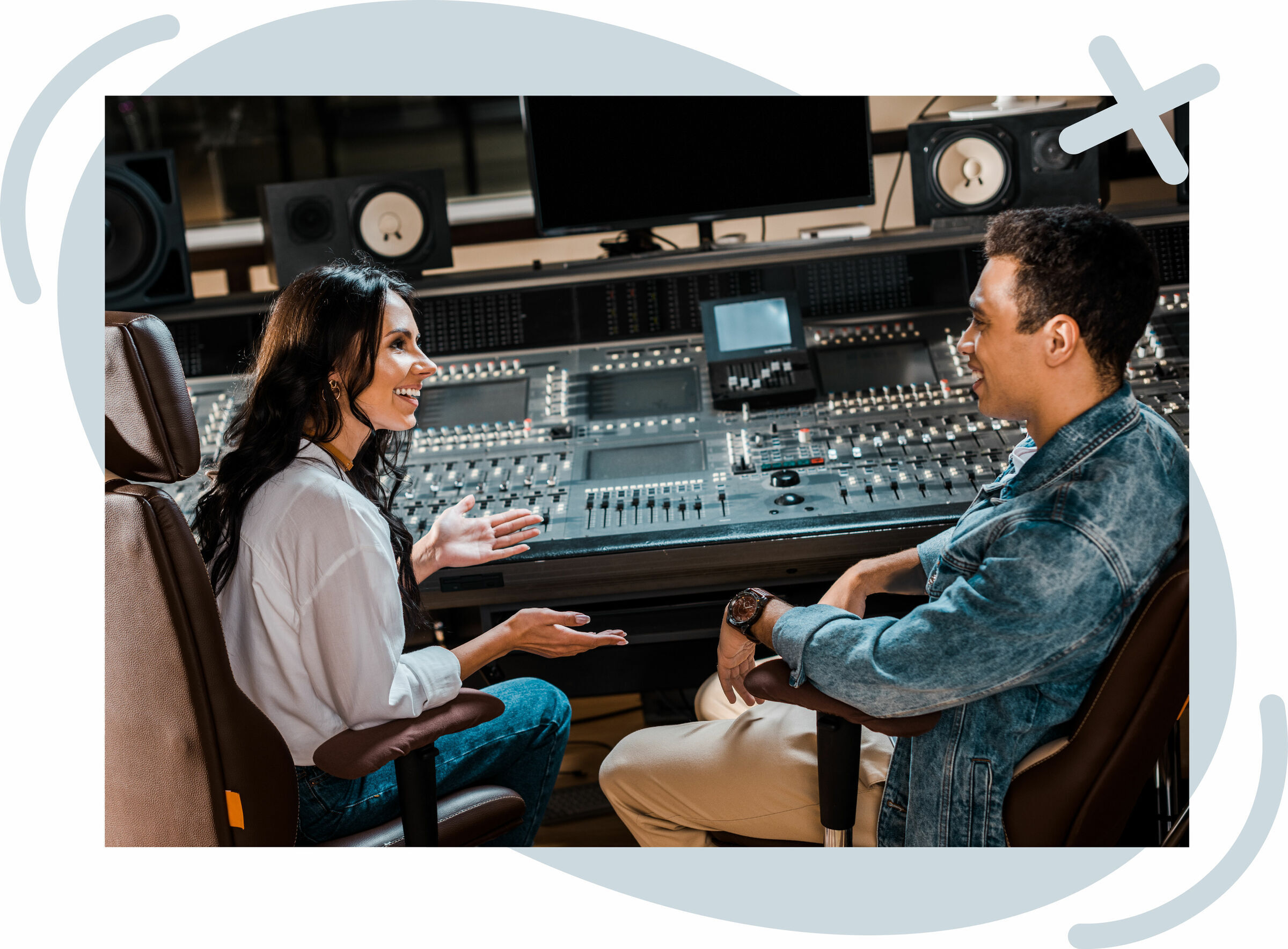 Two people talk and smile while sitting in front of a large audio mixing console in a recording studio.