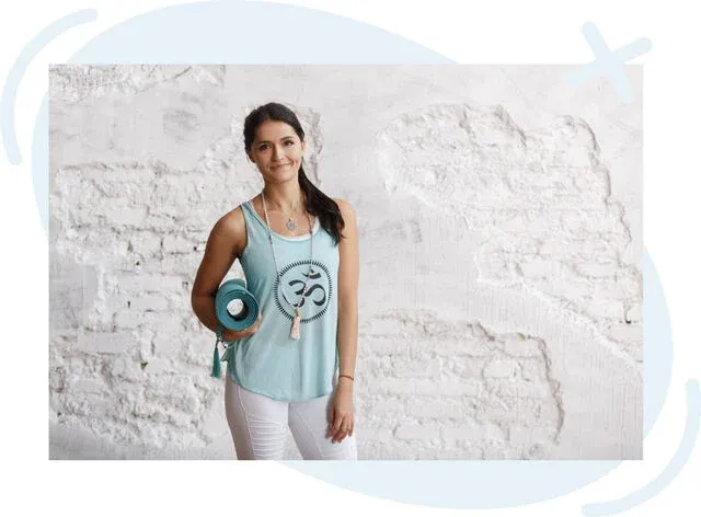 Young woman in a light blue tank top holding a rolled yoga mat, standing against a textured white brick wall and smiling softly at the camera.