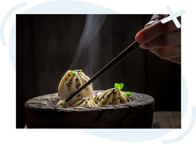 Hand using chopsticks to pick up a steaming dumpling garnished with sesame seeds and microgreens from a rustic wooden bowl.