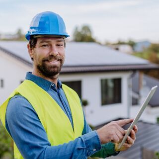 Smiling construction worker in a blue hard hat and neon safety vest uses a tablet while standing outside a modern house with rooftop solar panels.
