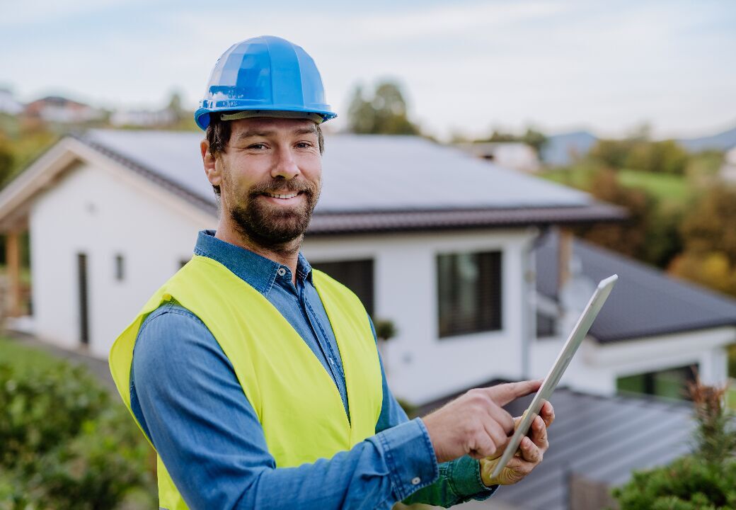 Smiling construction worker in a blue hard hat and neon safety vest uses a tablet while standing outside a modern house with rooftop solar panels.