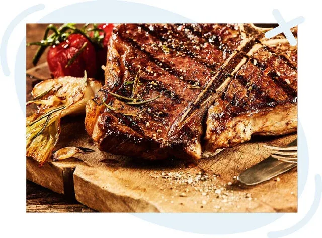Close-up of a grilled T-bone steak seasoned with coarse salt and herbs on a rustic wooden board, with roasted tomatoes and charred onions beside a fork and knife.