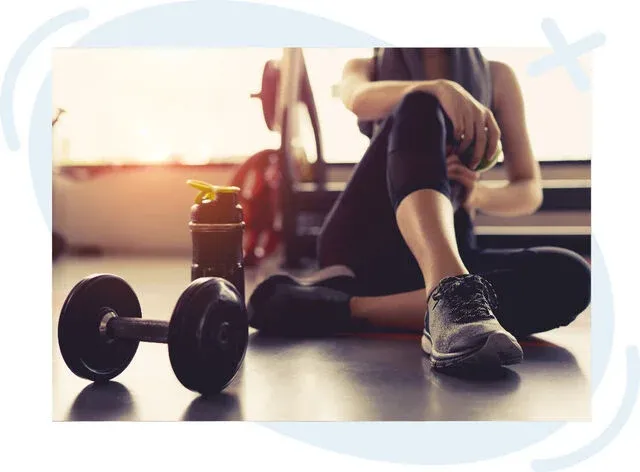 Person resting on a gym floor beside a dumbbell and a shaker bottle, with sunlight streaming through windows in the background.