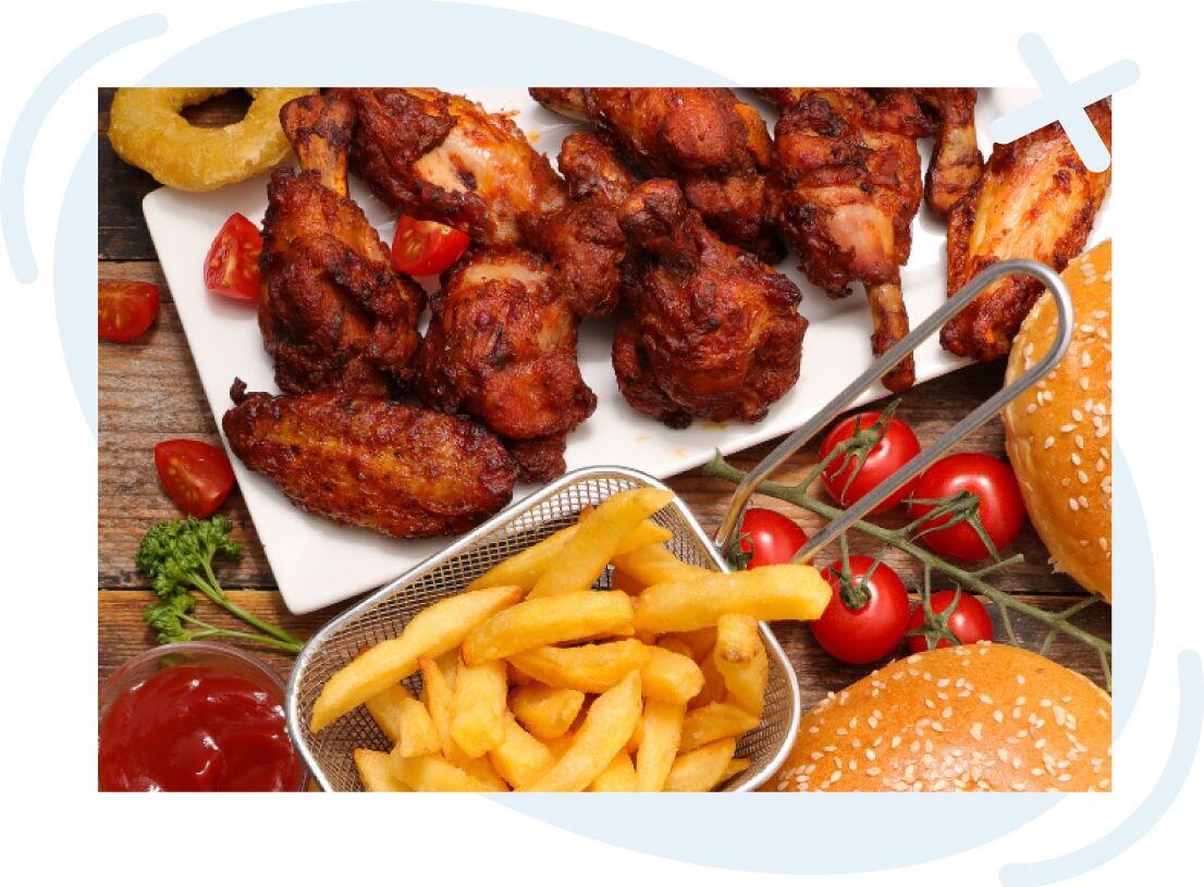 Close-up of a fried comfort food spread featuring crispy chicken wings and drumsticks on a white platter, a small basket of golden French fries, cherry tomatoes on the vine, sesame seed burger buns, and a dish of ketchup on a rustic wooden table.