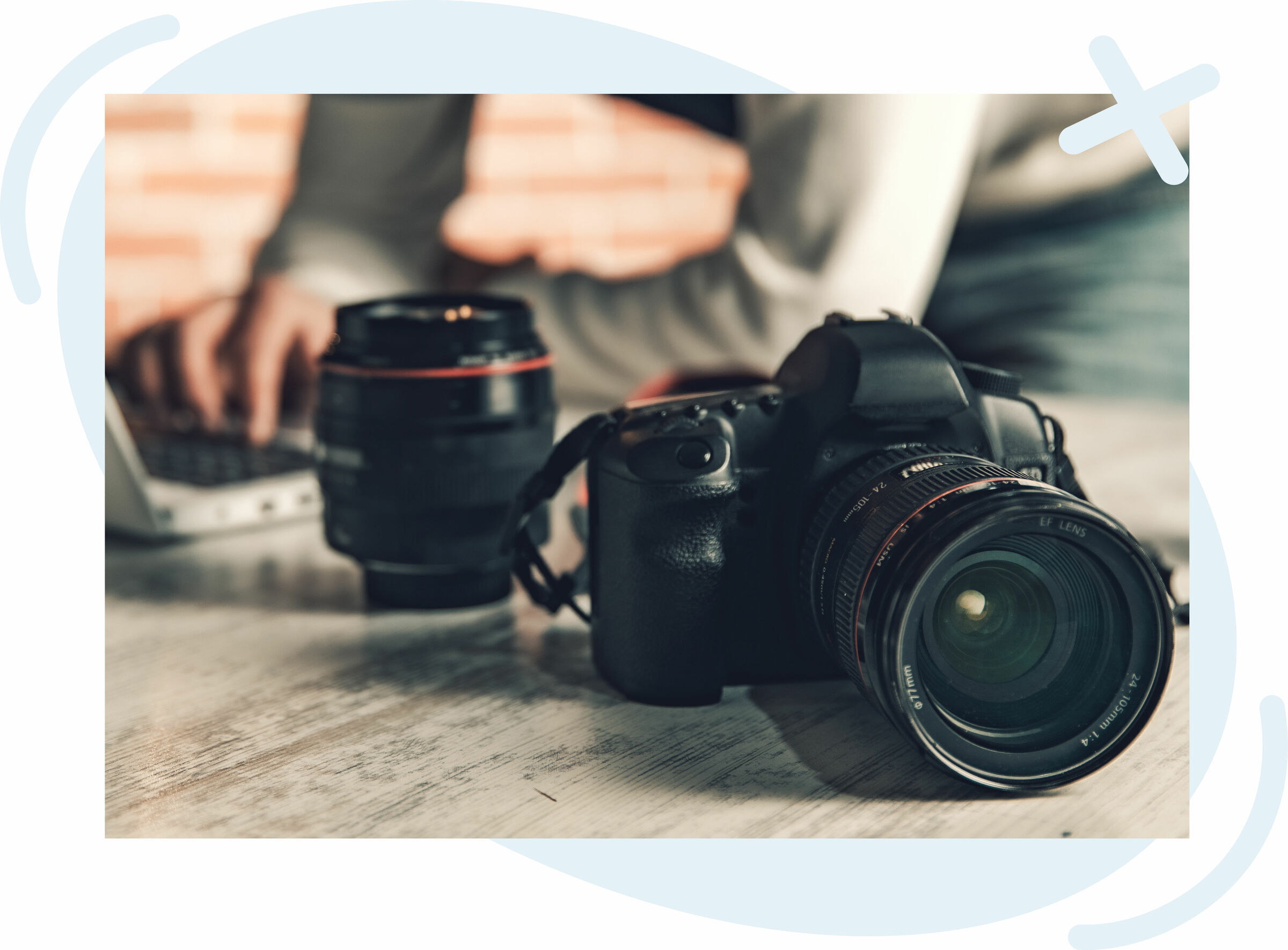 Close-up of a DSLR camera with a lens on a wooden table, next to an additional lens, while a person types on a laptop in the blurred background.