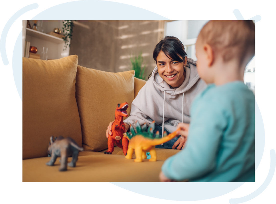 Smiling caregiver playing with a toddler using dinosaur toys on a mustard-colored couch in a cozy living room.