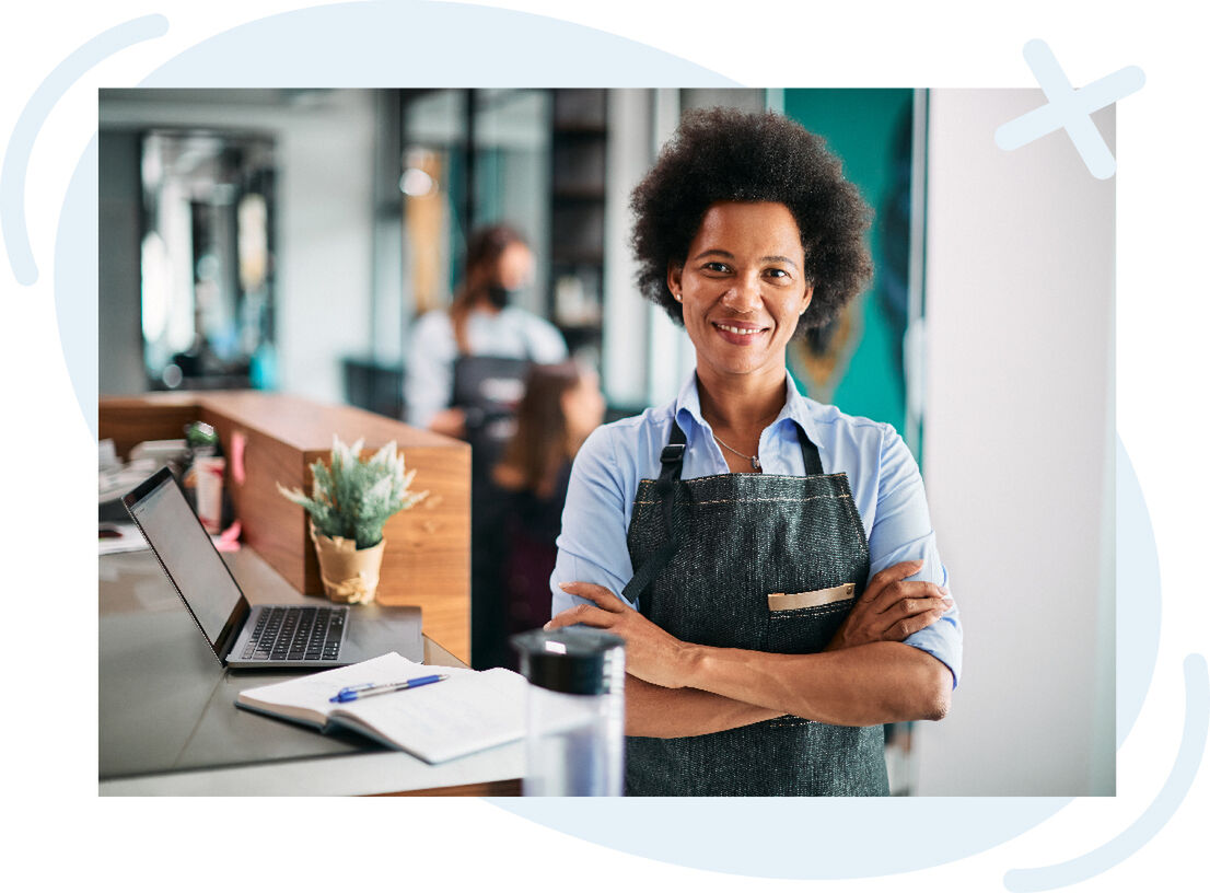 Confident shop worker in a denim apron stands with arms crossed at a modern reception counter, smiling toward the camera. A laptop, notebook with pen, small potted plant, and water bottle sit on the counter; blurred coworkers and shelves are visible in th