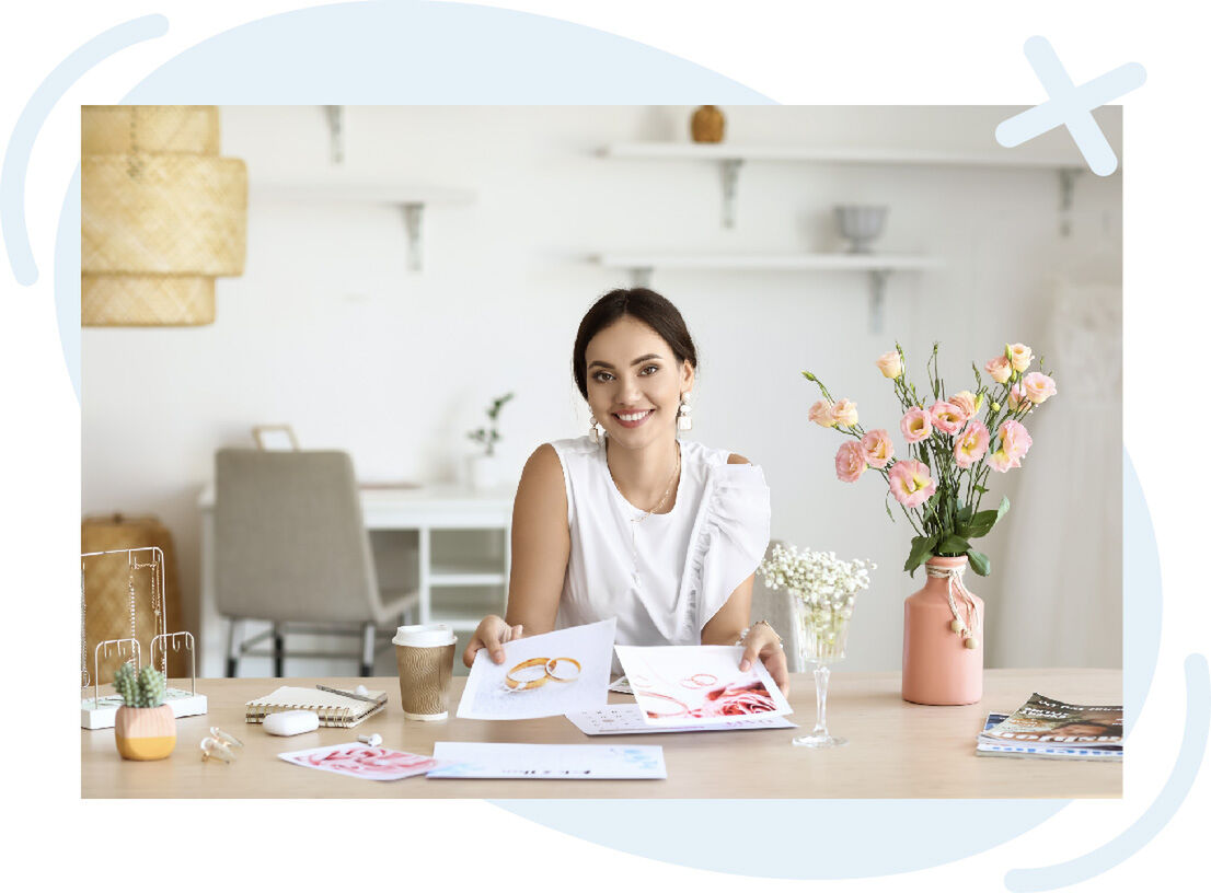 Smiling woman at a tidy desk holding design prints in a bright, modern studio with flowers and stationery around her.