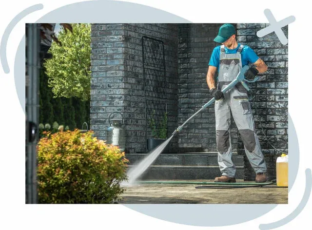 Worker pressure washing a brick patio outside a home, spraying water to clean the surface near steps and a porch.
