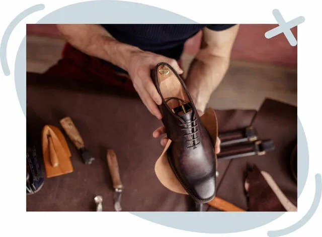 Artisan holding a handcrafted brown leather oxford shoe above a workbench with cobbler tools and leather pieces.