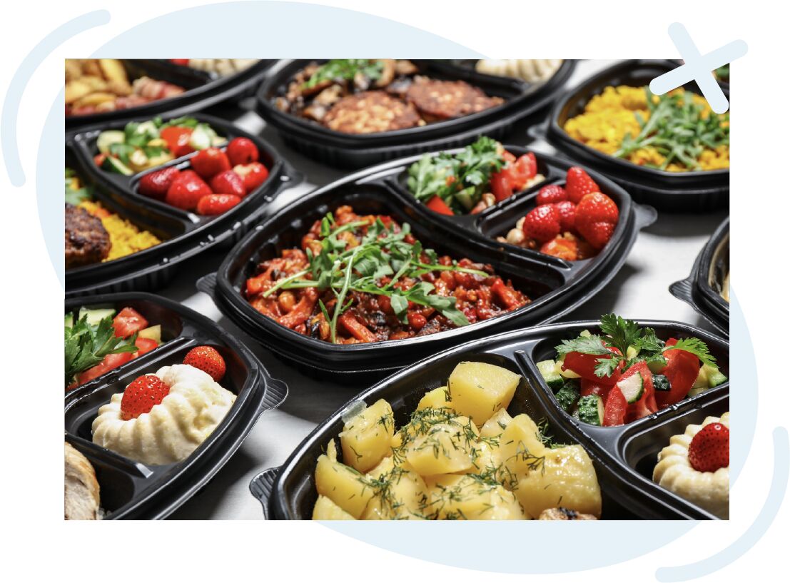 Assorted ready-to-eat meals in black compartmented containers with colorful vegetables, potatoes, strawberries, and garnishes.