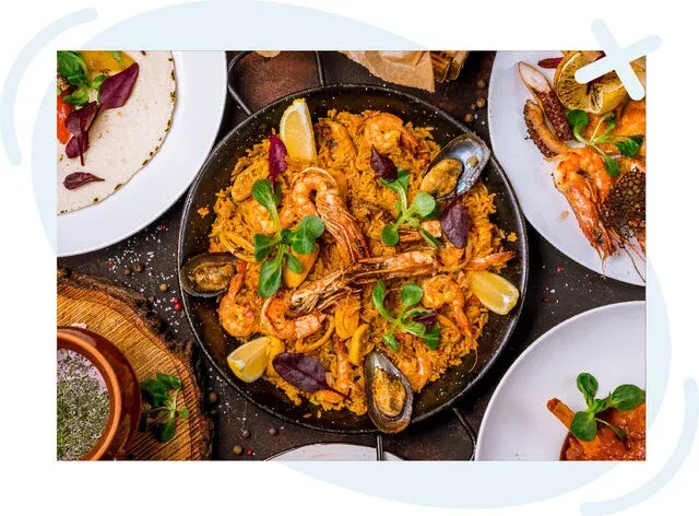Top-down view of a seafood paella in a black pan, garnished with lemon wedges and greens, surrounded by plates of seafood and sides on a dark tabletop.