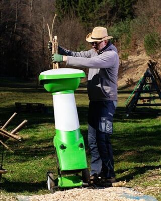 Person feeding branches into a green and white wood chipper in a grassy outdoor area, with a wheelbarrow full of sticks nearby.