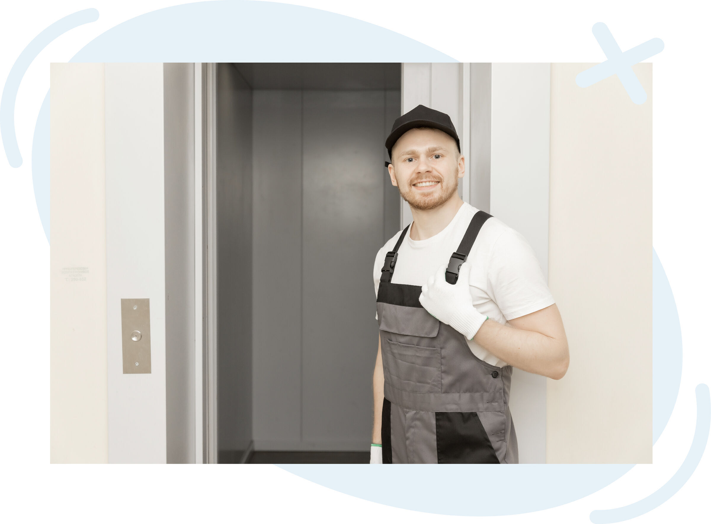Smiling technician in gray overalls and cap standing beside an open elevator doorway, gesturing toward the interior.