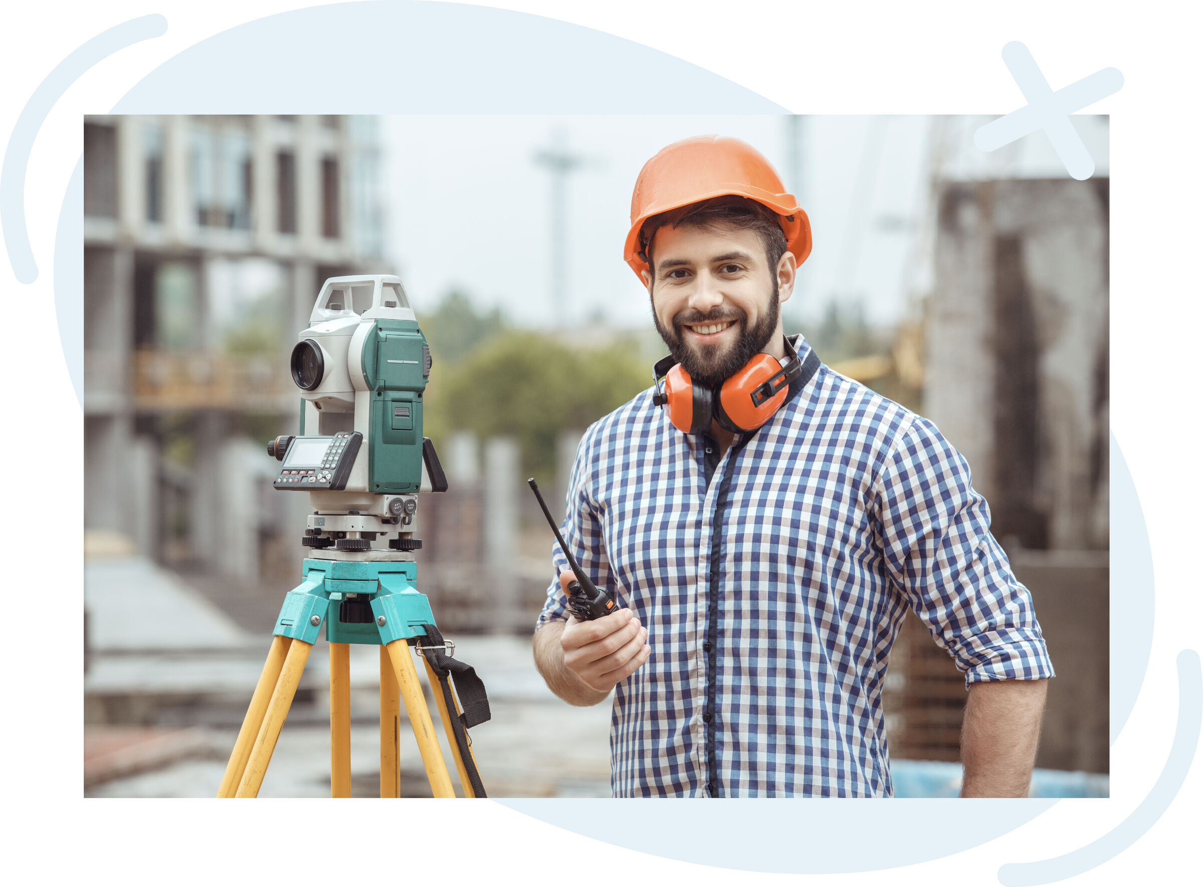 Surveyor at a construction site standing beside a total station on a tripod, wearing an orange hard hat and protective earmuffs around his neck, smiling and holding a walkie-talkie.