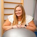 Smiling woman leaning on a large silver exercise ball in a bright indoor setting.