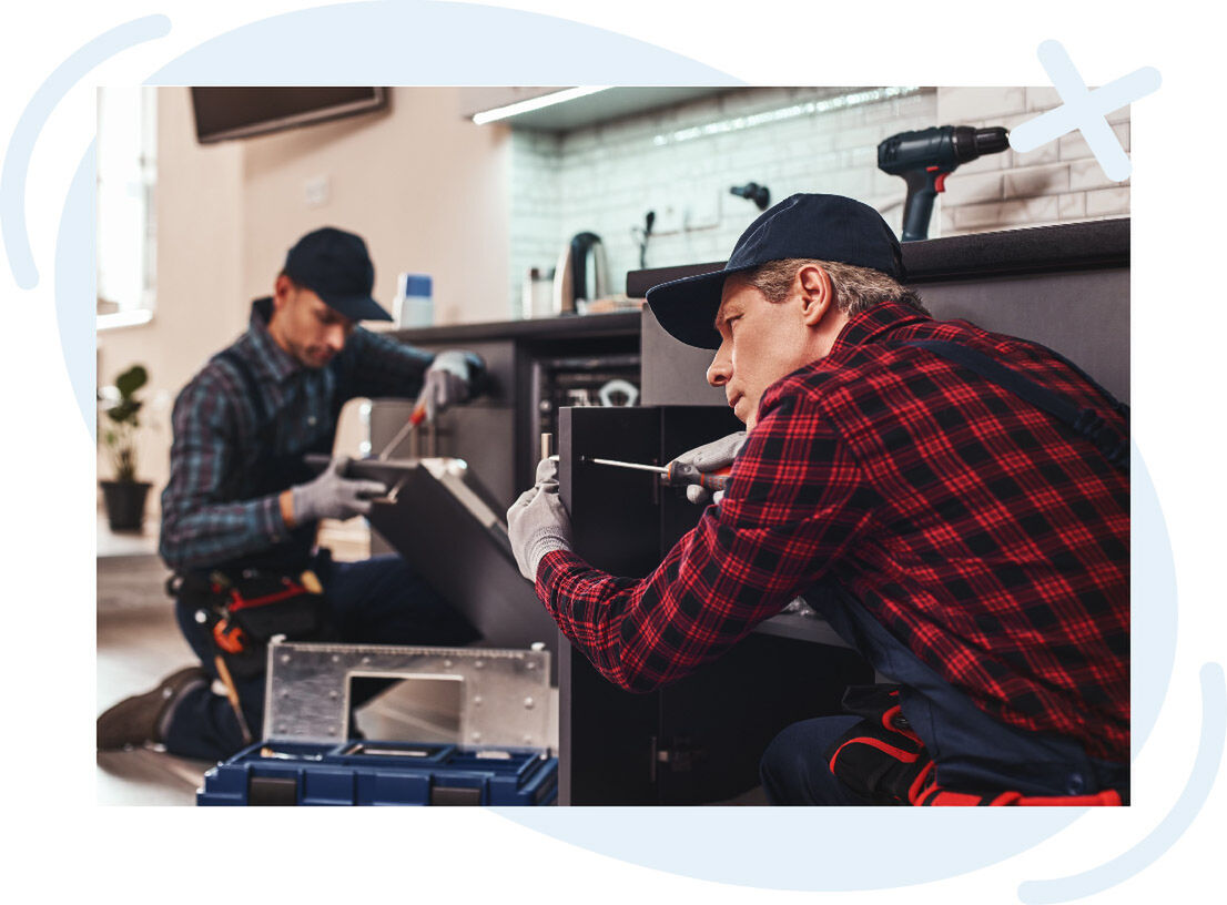 Two handymen in caps and work gloves assembling kitchen cabinets, using screwdrivers and tools on the floor of a modern kitchen.