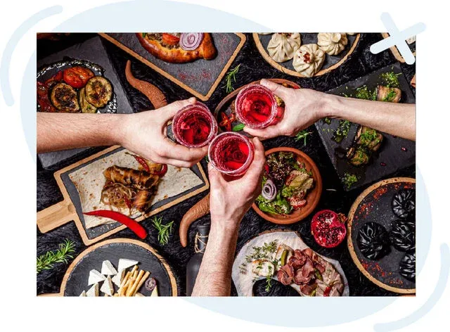 Top view of a celebratory toast over a spread of Georgian cuisine, with three hands clinking glasses of red drink amid assorted dishes on dark stoneware plates.