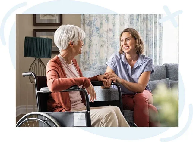 Younger woman sitting on a couch holding hands with an elderly woman in a wheelchair, smiling and talking in a cozy living room.