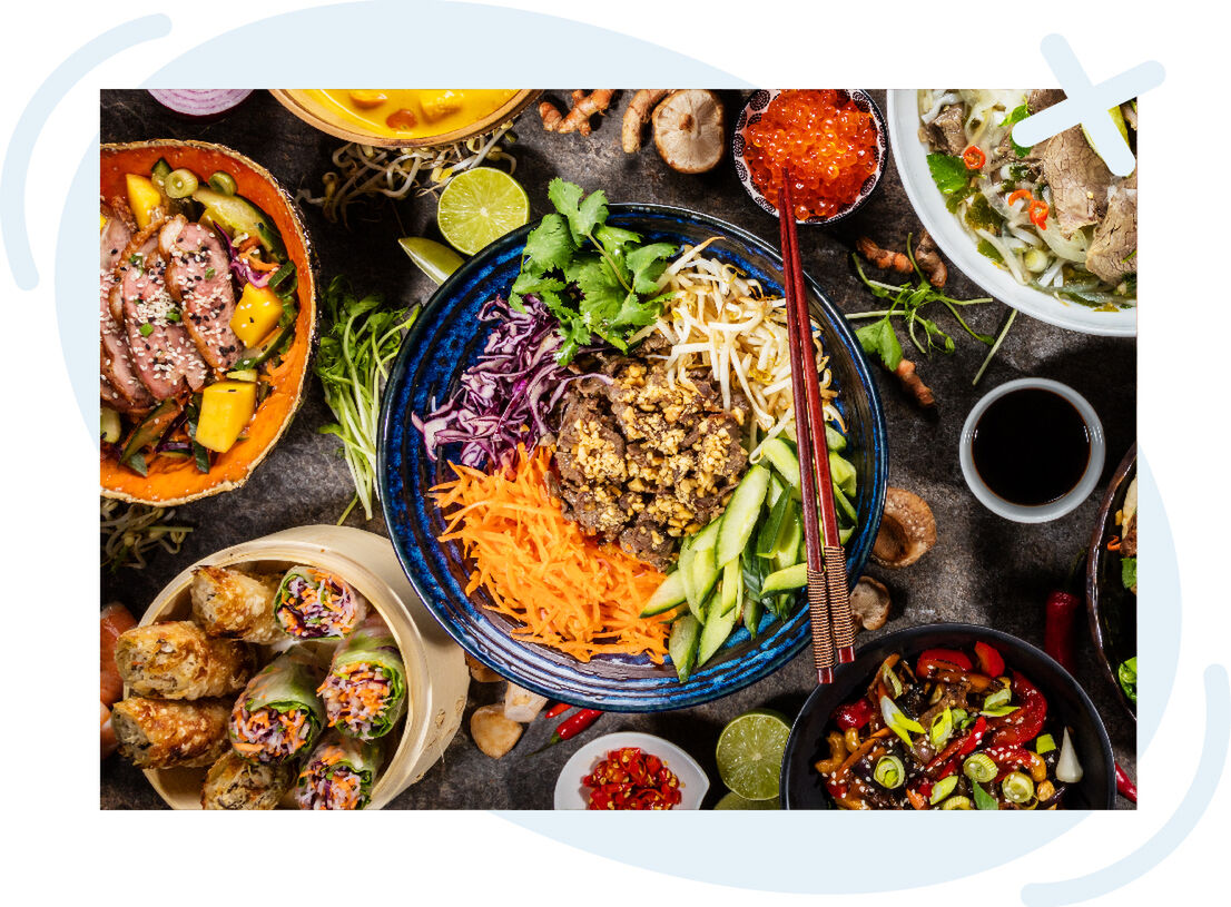 Colorful assortment of Asian-inspired dishes arranged on a table, centered on a vibrant noodle or rice bowl with shredded vegetables and chopsticks.