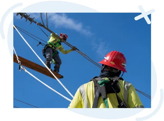 Two utility workers in safety gear repair overhead lines against a clear blue sky; one stands on a wooden beam high on the structure while the other observes from below, both wearing red helmets, harnesses, and reflective vests.
