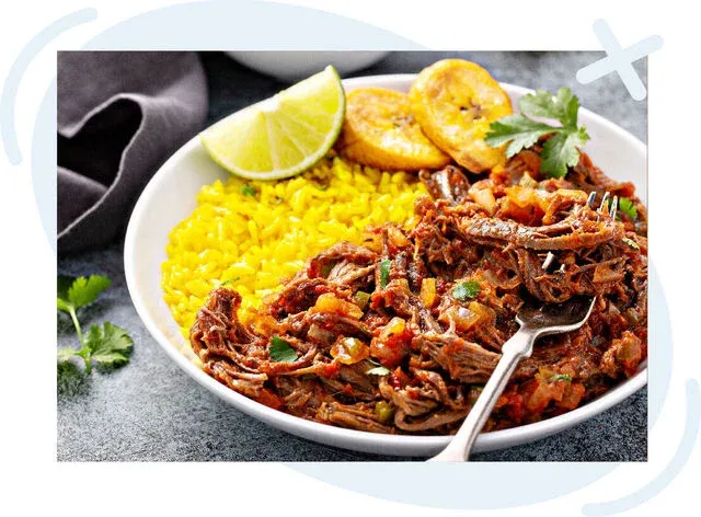Bowl of Cuban-style shredded beef with tomato and peppers served alongside bright yellow rice, fried plantain slices, and a lime wedge, garnished with cilantro.